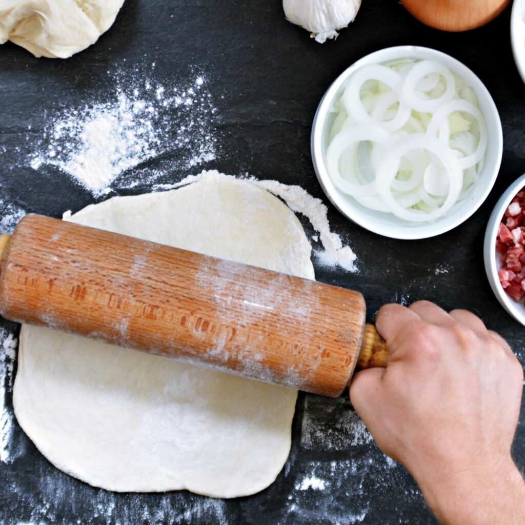 Flammkuchenteig wird mit einem Nudelholz ausgerollt.