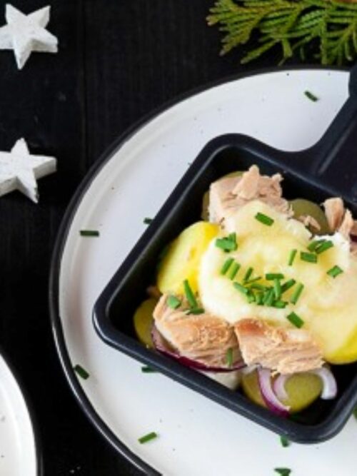 Raclette Pfännchen mit Kartoffeln und Thunfisch wird auf einem weißen Teller mit Schnittlauch gezeigt.