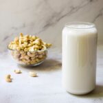 Cashewmilch wird in einem Glas vor einem Marmor-Hintergrund mit einer Schale Cashews gezeigt.