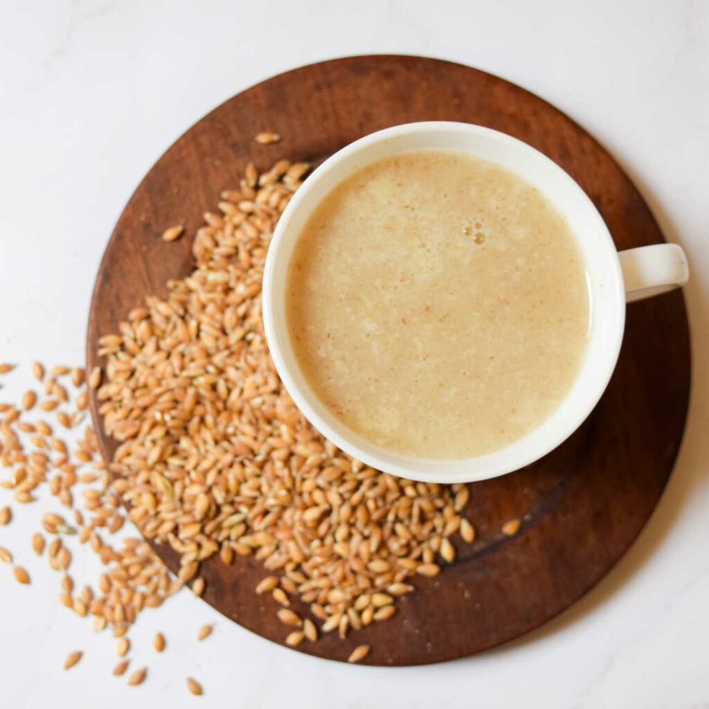 Dinkelmilch wird in einer weißen Tasse auf einem hölzernen Teller mit ein paar Dinkelkörnern von oben gezeigt.