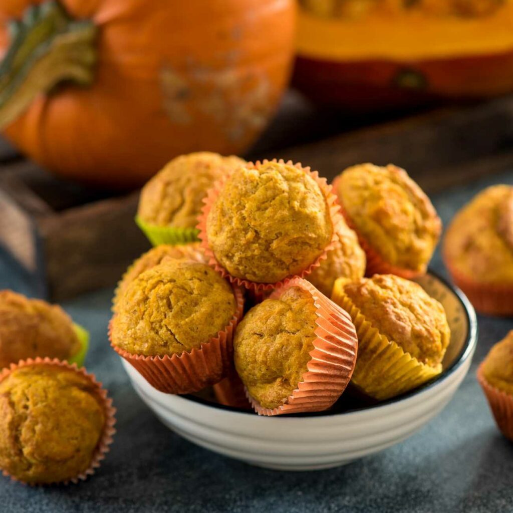 Kürbis-Muffins werden gestapelt in einer Schale mit Kürbissen im Hintergrund gezeigt.