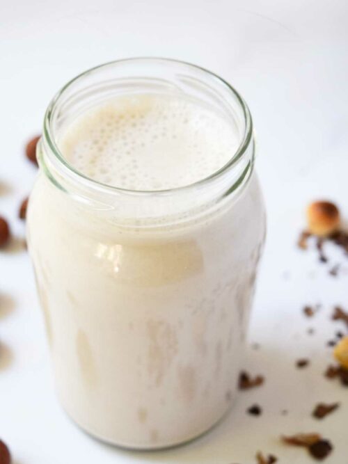 Milch aus gerösteten Haselnüssen wird in einer kleinen Glasflasche von schräg oben und von ein paar Haselnüssen umgeben gezeigt.
