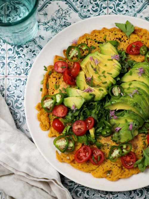 Veganes Omelett wird mit Salat und Avocado auf einem Teller auf bunten Fliesen gezeigt.