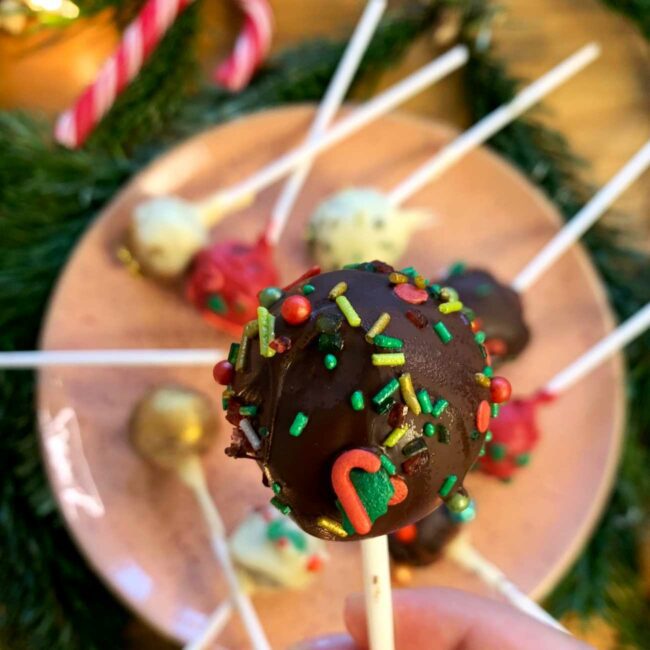 Weihnachtliche Cake Pops werden unscharf auf einem Teller gezeigt, während einer in den Vordergrund gehalten wird.