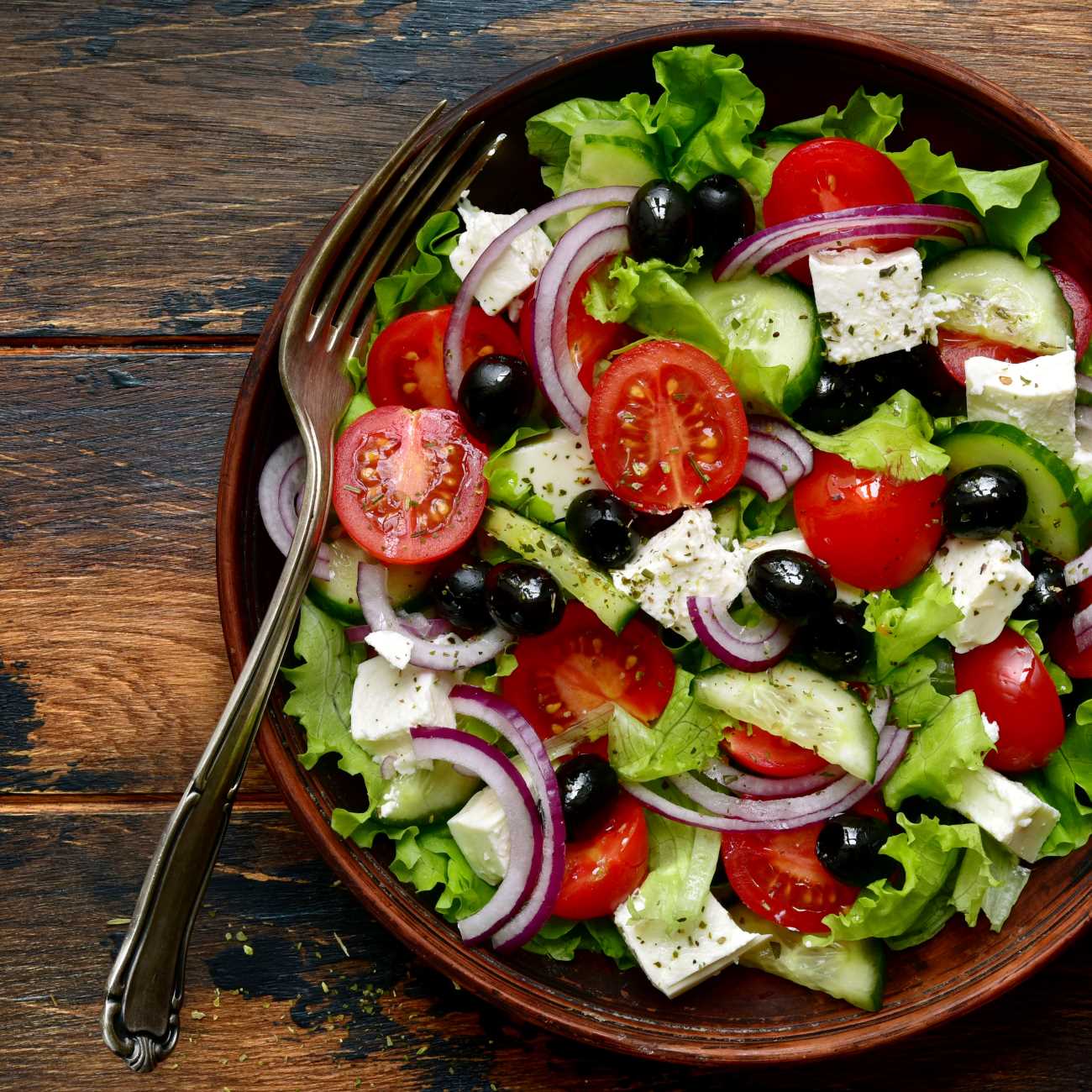 Griechischer Salat wird in einer braunen Schüssel auf Holzuntergrund gezeigt.