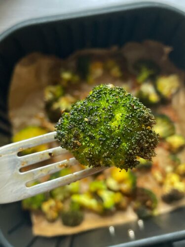 Brokkoli aus der Heißluftfritteuse wird auf einer Gabel nah an die Kamera gehalten. Die Pfanne der Heißluftfritteuse ist darunter unscharf zu sehen.